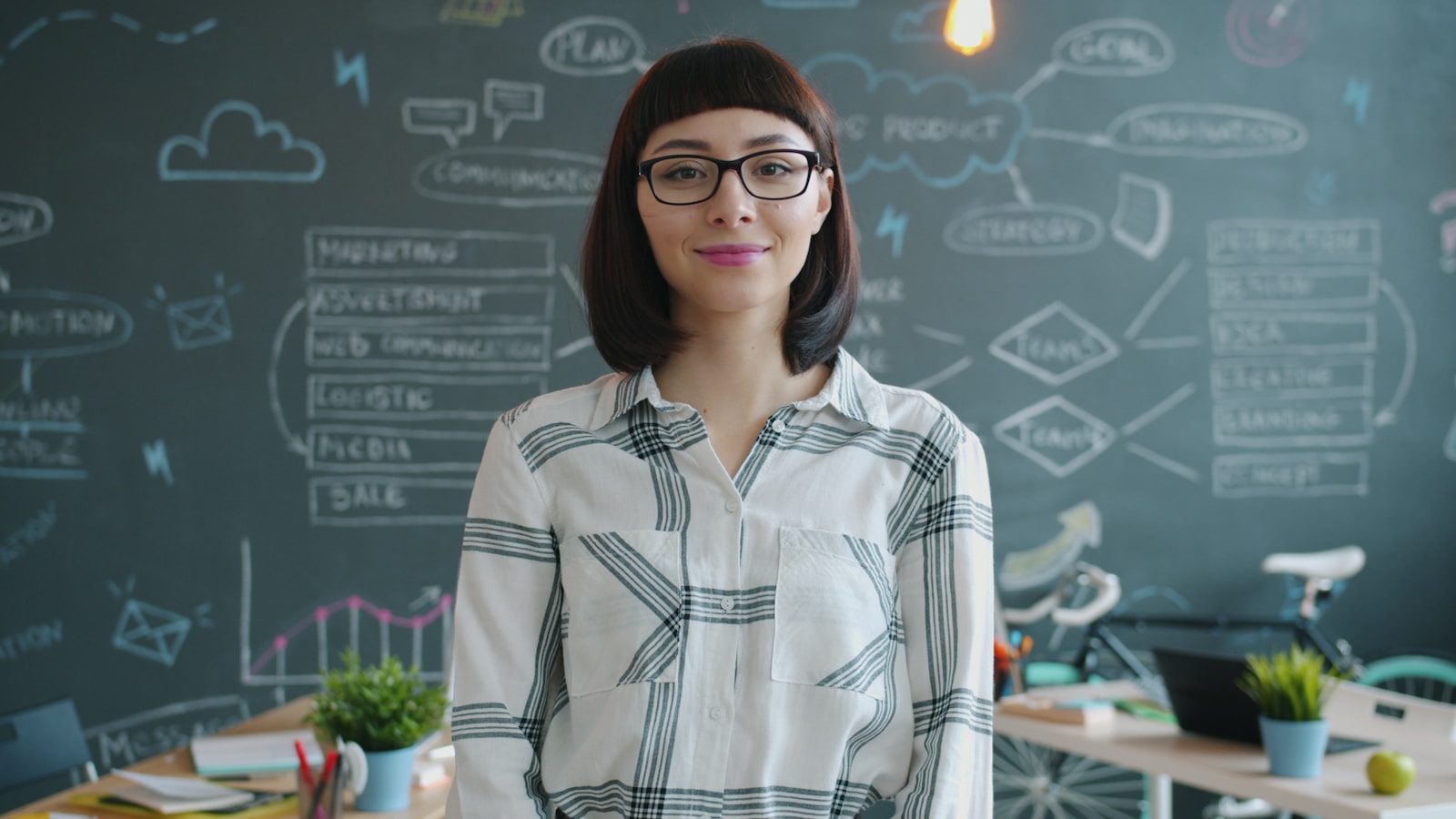 Woman with glasses smiles in front of chalkboard.